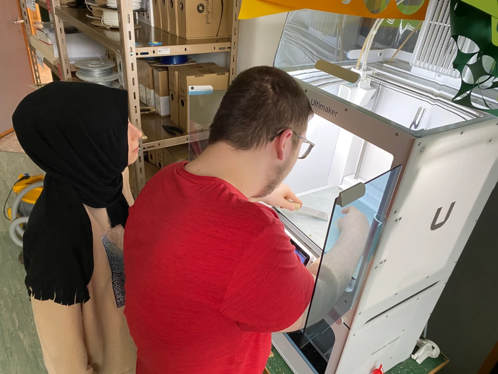Zwei Schüler:innen vor einem 3D-Drucker. Schüler arbeitet mit Spatel auf dem Druckbett im 3D-Drucker. Im Hintergrund ein Regal mit Filamentspulen.