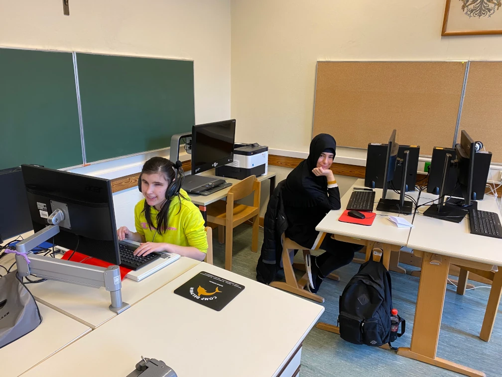 Zwei Schülerinnen an ihrem Computer-Arbeitsplatz. Eine Schülerin trägt ein Headset und sitzt vor einem Monitor, Tastatur und Braillezeile. Die zweite Schülerin trägt ein Kopftuch und sitzt vor einem Monitor, Tastatur und Maus. Weitere Computer-Arbeitsplätze, ein Drucker sowie eine Tafel und Pinnwand sind abgebildet.