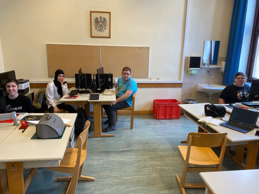 Vier Schüler:innen sitzen an ihren Computerarbeitsplätzen und lächeln in die Kamera. Die Computerarbeitsplätze sind in drei Tischgruppen angeordnet. In der Mitte stehen zwei leere Sessel. An der Wand im Hintergrund befindet sich eine Pinnwand, ein Bild vom Bundeswappen und eine Waschgelegenheit.