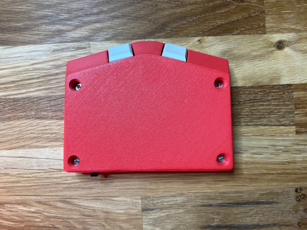 Die Rückseite der mobilen Braille-Tastatur Metabraille. Das Tastaturgehäuse ist rot und aus Kunststoff (125 mm x 95 mm x 25 mm). Die Gehäuseform ist rechteckig, mit einer flachen, trapezförmigen Erweiterung auf der Längsseite. Zwei weiße Tasten liegen auf der trapezförmigen Erweiterung in einem Abstand von ca. 20 mm. Vier versenkte Schrauben befinden sich an den Ecken des Gehäuses.